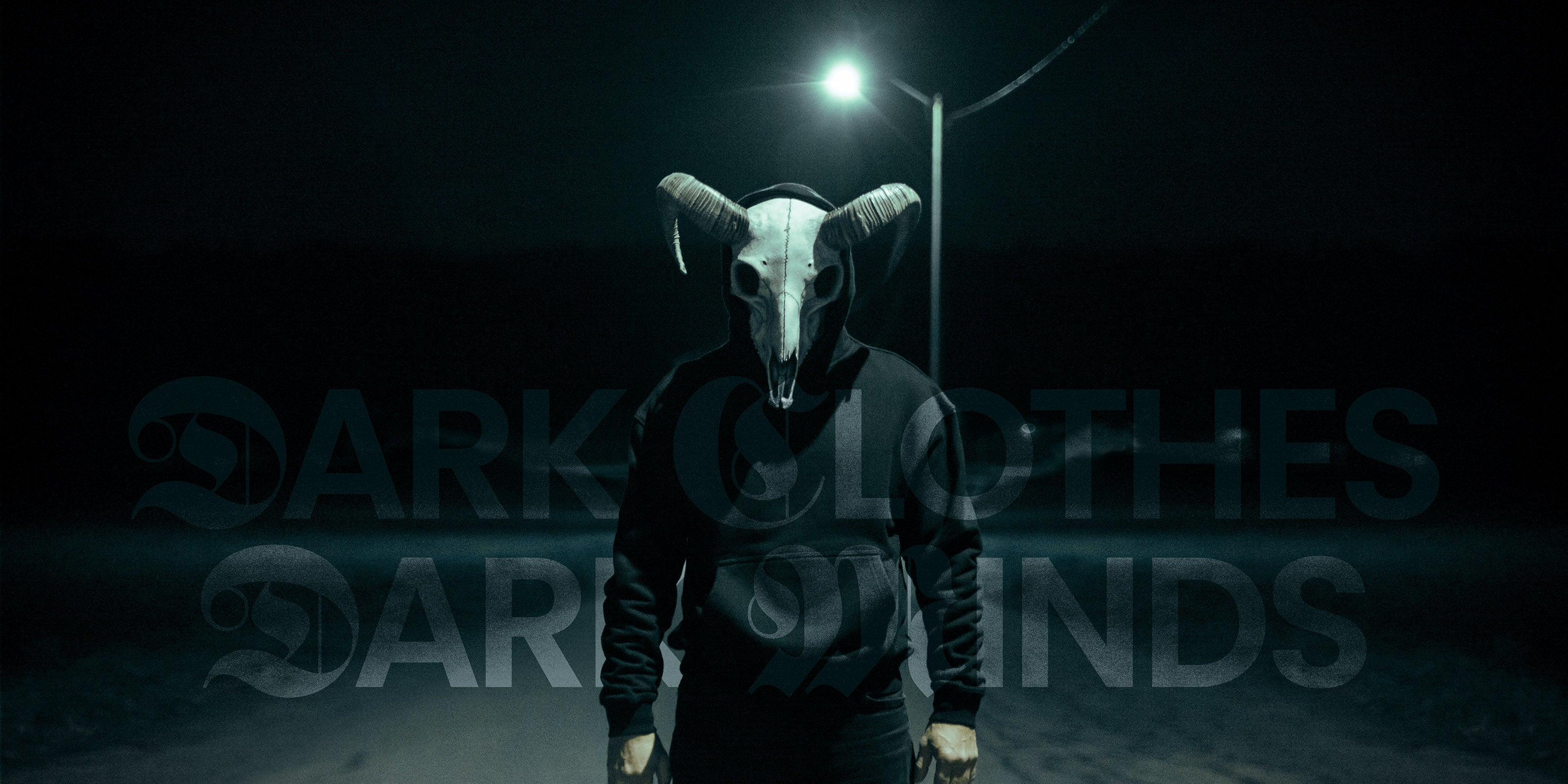 Person wearing a goat skull mask and a hoodie in a dark, urban setting with streetlights.