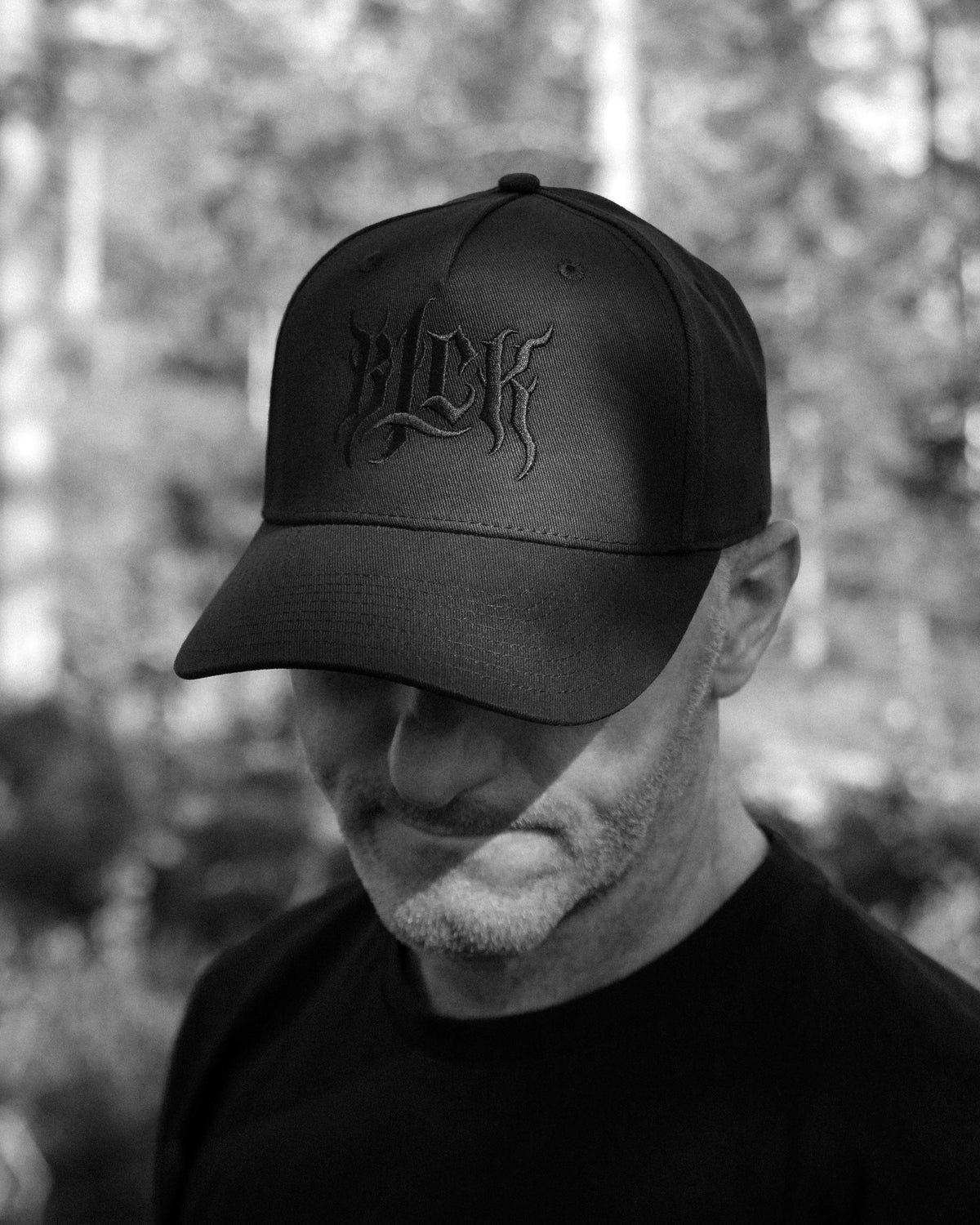Person wearing a black cap with an embroidered BLCK logo, in a forest background