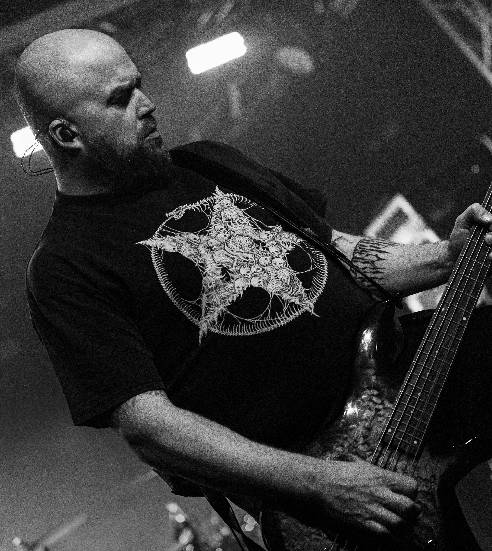 Person playing bass guitar with a detailed pentagram design on a black t-shirt, against a blurred stage background.